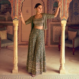 May include: A green floral print three-piece outfit, including a crop top, jacket, and wide-leg pants. The outfit features intricate embroidery on the top and jacket. The model is standing in a room with ornate architectural details.