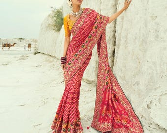 May include: A woman wearing a vibrant pink saree with intricate embroidery and a yellow blouse. The saree features detailed patterns and a flowing design, with a decorative border. The background shows a desert landscape.