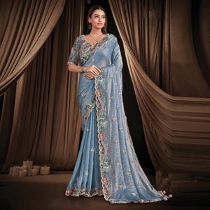 Könnte beinhalten: Ein hellblaues Sari mit aufwendiger Stickerei und einem detaillierten Saum. Das Sari hat eine passende Bluse mit verzierten Ärmeln. Das Model trägt eine Halskette und Ohrringe. Der Hintergrund umfasst braune Vorhänge und brennende Kerzen.