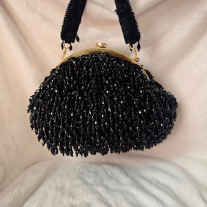 May include: A vintage black beaded handbag with a gold-tone clasp and black velvet handles. The bag is covered in small, shiny black beads, creating a textured surface. The handles are attached with small gold-tone rings.