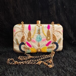 Puede incluir: Clutch rectangular marfil con herrajes dorados y una cadena dorada. El clutch está adornado con bordados coloridos, incluyendo detalles en rosa, azul, amarillo y dorado. La palabra "CLUTCHES" está impresa en la parte delantera.