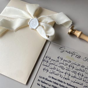 Może przedstawiać: Zestaw zaproszeń w kolorze kości słoniowej z kremową wstążką i białą pieczęcią woskową. Przezroczysta akrylowa karta wyświetla kaligrafię i tekst. Obecny jest również drewniany stempel.