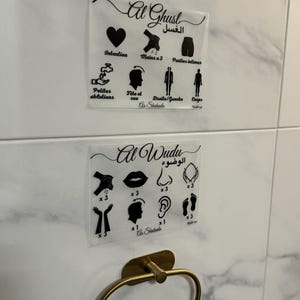 Może przedstawiać: Dwie przezroczyste akrylowe instrukcje z czarnymi ilustracjami i tekstem, jedna z napisem "Al Ghusl", a druga "Al Wudu". Poniżej, złoty wieszak na ręczniki jest zamontowany na białej ścianie z płytek z wzorem marmuru.