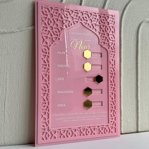 Może przedstawiać: Różowy akrylowy tracker modlitewny z geometrycznymi wycięciami. Tekst "LES CINQ PRIERES" i arabski skrypt u góry, ze złotym napisem "Nour". Posiada pięć sekcji ze złotymi i czarnymi sześciokątnymi znacznikami oraz tekstem "A FAIRE" i "OK".