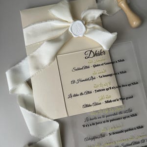 Może przedstawiać: Biała koperta z białą satynową wstążką zawiązaną w kokardę i białą pieczęcią woskową. Koperta leży na przezroczystej plastikowej kartce z tekstem arabskim i tłumaczeniami na język francuski. Tekst brzmi "Dhikr" i zawiera takie zwroty jak "SubhanAllah", "Al Hamdulilah", "La ilaha illa Allah", "Allahu Akbar", "La hawla wa la quwatah illa bil Lah", "Astaghfiru Lah" i "SubhanAllahi wa bi Hamdiki, SubhanAllahi At".