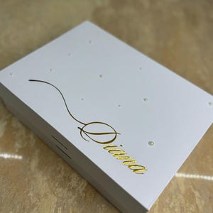 Może przedstawiać: Białe pudełko prezentowe z złotym napisem "Diana" i małymi białymi perełkowymi ozdobami.