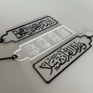 Może przedstawiać: Trzy prostokątne, przezroczyste akrylowe zakładki do książek z czarno-białą kaligrafią arabską. Każda zakładka ma unikalny wzór i jest przymocowana do czarnego sznurka. Zakładki ułożone są na jasnoszarej powierzchni.