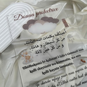 Może przedstawiać: Przezroczysty znak akrylowy z brązowym napisem "Douaa protectrice". Znak przedstawia białą i szarą chmurę z małym pluszowym misiem trzymającym balony. Znak zawiera również tekst arabski.