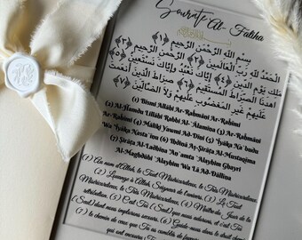 May include: A white framed print with the Arabic text of the first chapter of the Quran, Surah Al-Fatiha, in black script. The text is translated into English below the Arabic text. The print is on a light gray background.