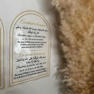 Może przedstawiać: Dwa białe znaki z złotymi obramowaniami i arabskim tekstem w kolorze czarnym. Znaki są zamontowane na białej ścianie. Tekst na górnym znaku brzmi "En entrant à la maison", a tekst na dolnym znaku brzmi "En sortant de la maison".