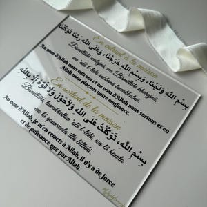 Może przedstawiać: Prostokątna, przezroczysta płyta akrylowa ze złotym i czarnym tekstem w języku arabskim i angielskim. Tekst zawiera zwroty dotyczące wchodzenia i wychodzenia z domu. Biała wstążka jest ułożona na górze.