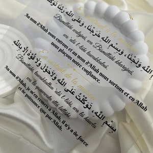 Może przedstawiać: Biały znak akrylowy z pozłacanymi literami przedstawiający islamską modlitwę w języku arabskim i francuskim. Modlitwa dotyczy wchodzenia i wychodzenia z domu z błogosławieństwem Boga. Znak jest ozdobiony białym koralikowym łańcuszkiem.