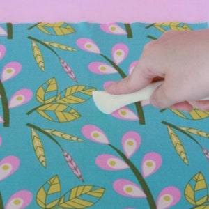 May include: A white plastic tool with a rounded edge is being used to smooth out a piece of fabric with a blue background and a repeating pattern of pink and yellow leaves.