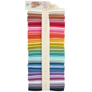 May include: A stack of fabric squares in a rainbow of colors, secured with a cream-colored ribbon. The ribbon is printed with the words "Tilda Collection Solids." A tag at the top features floral designs and the word "Solids."
