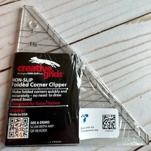 May include: A clear plastic triangle ruler with a black and white packaging featuring a fox logo and the text "creative. grids The Original NON-SLIP Non-Slip Folded Corner Clipper Make folded corners quickly and accurately - no need to draw pencil lines! Designed by Susan Nelson CGRFCC Made in USA SEE A DEMO SCAN WITH ANY QR READER Scan with the Transparency app CGRFCC".