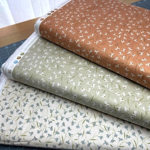 May include: Three rolls of fabric with floral patterns. The top roll is a warm brown with white and brown floral sprigs. The middle roll is light green with white floral sprigs. The bottom roll is off-white with blue floral sprigs.