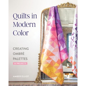 May include: A book cover titled "Quilts in Modern Color" with the subtitle "Creating Ombré Palettes" and author Amber Elliot. The image shows a mirror reflecting colorful geometric quilts in shades of orange, pink, and purple.