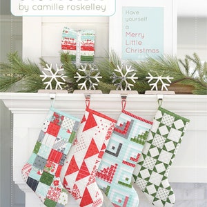 May include: Four quilted Christmas stockings hanging on a white mantel with a sign that reads "Have yourself a Merry Little Christmas". The stockings are made with red, green, blue, and white fabrics. The sign is on a white background with blue text.