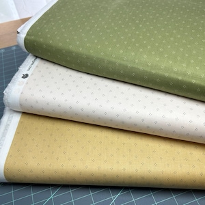 May include: Three stacked fabric bolts in shades of green, cream, and gold. Each fabric has a repeating pattern of small, dark square shapes. The edges of the fabric are white.