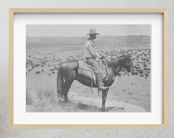 May include: A black and white photograph of a cowboy on horseback riding through a herd of cattle. The cowboy is wearing a hat and a long coat. The photograph is framed in a wooden frame and hanging on a wall.