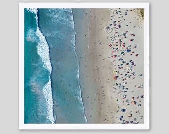 May include: An aerial view of a crowded beach with blue ocean waves crashing on the shore. The beach is covered in colorful umbrellas and people.