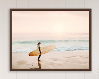 May include: A woman in a hat stands on a sandy beach holding a surfboard. The ocean is behind her and the sky is a soft pink color.