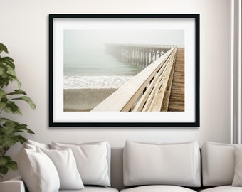 May include: A photograph of a wooden pier extending out over a foggy ocean. The pier is made of weathered wood and has white railings. The ocean is a light blue color and the fog is a soft white.