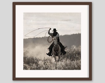 May include: Sepia-toned framed print of a cowboy on horseback, roping. The cowboy, wearing a hat and vest, is in motion, with the rope extended. The horse is running through a field of tall grass, creating a sense of action and the wild west.