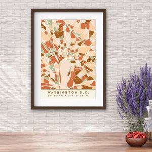 May include: Framed art print of a map of Washington D.C. in a geometric style. The artwork features a color palette of beige, brown, and green. The city name and coordinates are printed at the bottom. The frame is dark brown.