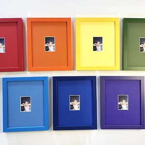 May include: Seven colorful picture frames in red, orange, yellow, green, blue, dark blue, and purple. Each frame has a small, rectangular photograph with a white mat. The frames are mounted on a white wall.