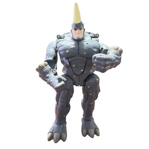 May include: A gray action figure of a muscular humanoid character with a yellow horn on its head. The figure has a textured, armored appearance with gray and light gray accents. The figure is posed with its fists clenched.