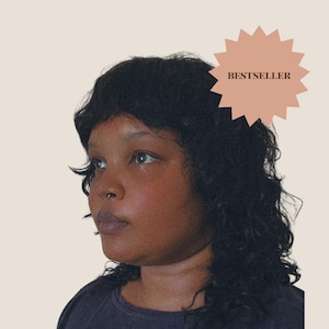 May include: A person with dark, shoulder-length wavy hair and a small silver septum ring in their nose. The person is wearing a dark-colored shirt. A peach-colored starburst graphic in the upper right corner contains the text BESTSELLER.