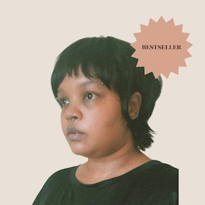May include: A person with a short black layered haircut, a septum piercing, and an eyebrow piercing. The person is wearing a black shirt. A light pink starburst graphic in the upper right corner contains the dark brown text BESTSELLER.