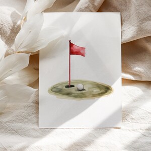 Puede incluir: Ilustraci&oacute;n en acuarela de una escena de golf. Una bandera roja ondea sobre un poste, marcando el hoyo. Una pelota de golf blanca descansa cerca del hoyo sobre un &aacute;rea verde y cubierta de hierba. La obra de arte est&aacute; sobre un fondo blanco.
