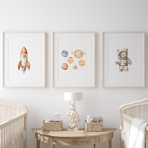 May include: Three framed watercolour prints with a space theme. The prints feature a rocket, planets, and a teddy bear astronaut. The prints are displayed on a white wall above a wooden table with a lamp and two cots.