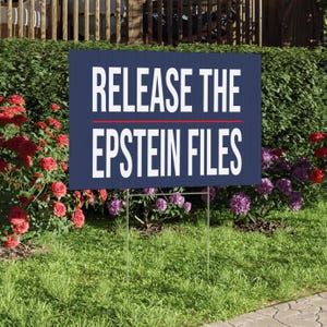 May include: A navy blue sign with the white text "RELEASE THE EPSTEIN FILES", separated by a red line. The sign is supported by a metal wire frame and placed in a grassy area with flowers and foliage in the background.
