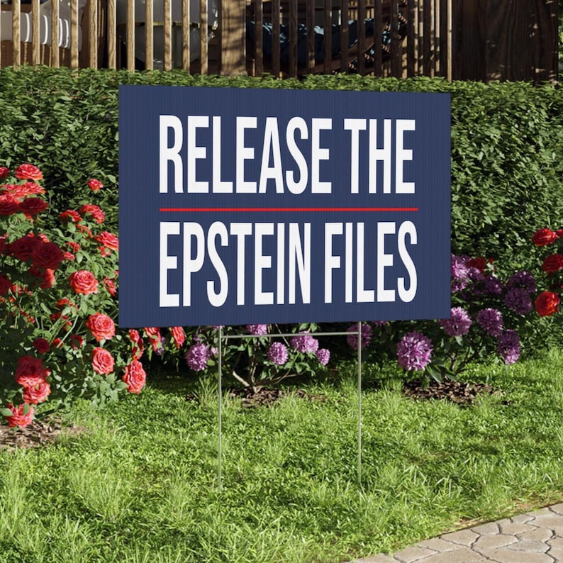 May include: A navy blue sign with the white text "RELEASE THE EPSTEIN FILES", separated by a red line. The sign is supported by a metal wire frame and placed in a grassy area with flowers and foliage in the background.