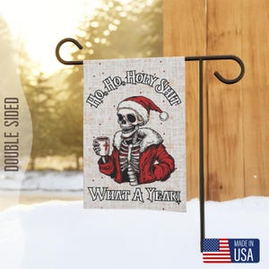 Könnte beinhalten: Eine doppelseitige Gartenflagge mit einem Skelett, das einen Weihnachtsmann-Hut trägt und eine Tasse hält. Die Flagge hat einen weißen Hintergrund mit rotem und schwarzem Text, der "Ho Ho Holy Shit What A Year" lautet.
