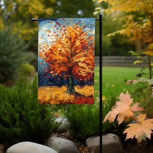 Puede incluir: Una bandera de jardín con una pintura de un árbol con hojas naranjas, amarillas y rojas. El árbol está sobre un fondo azul con algunas nubes blancas.