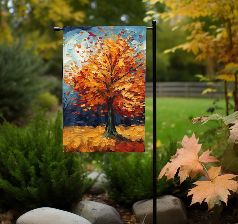 May include: A garden flag with a painting of a tree with orange, yellow, and red leaves. The tree is set against a blue background with a few white clouds.