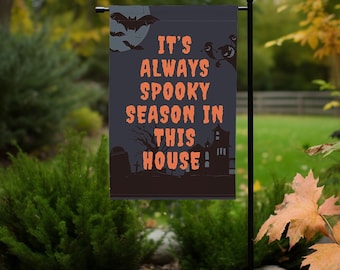 Puede incluir: Una bandera de jardín de Halloween negra y naranja con el texto "It's always spooky season in this house!" La bandera presenta una silueta de una casa, un árbol, un cementerio, murciélagos y fantasmas.