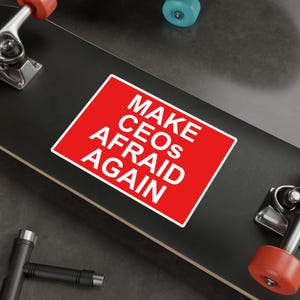 May include: A red sticker with white text that reads "MAKE CEOS AFRAID AGAIN" is placed on a black skateboard.