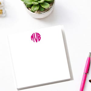 May include: A white notepad with a fuchsia monogram, a succulent plant in a white pot, a pink pen, and tortoiseshell glasses are arranged on a white surface. A gold stapler and a light blue book are also visible.