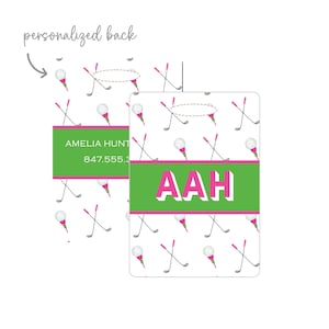 May include: A rectangular luggage tag featuring a white background with a repeating pattern of golf balls on pink tees and crossed golf clubs. A horizontal green band with pink borders displays the white capital letters AAH in the center.