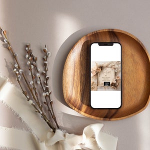 May include: A smartphone displaying a photo of a wooden sign with the text "It's a Girl!" and a baby ultrasound image. The sign is decorated with dried flowers and greenery. The phone is resting in a wooden bowl on a light brown surface.
