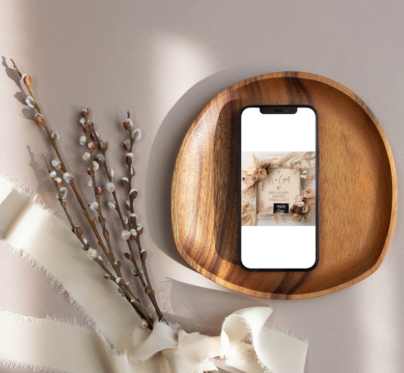 May include: A smartphone displaying a photo of a wooden sign with the text "It's a Girl!" and a baby ultrasound image. The sign is decorated with dried flowers and greenery. The phone is resting in a wooden bowl on a light brown surface.