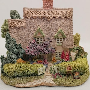 May include: A detailed miniature cottage with a thatched roof, brick chimney, and a garden. The cottage has a stone path, white picket fence, and an easel with a painting. The scene is surrounded by greenery and colourful flowers, evoking a quaint, rural setting.
