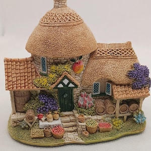 May include: A detailed miniature cottage with a thatched roof, stone walls, and a green door. The cottage is adorned with barrels, baskets of fruit, and grape vines. A peach hangs from the roof. The overall theme is a charming village scene.