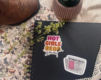 May include: A black spiral-bound notebook featuring two stickers. One sticker reads HOT GIRLS READ smut in orange, yellow, and pink text. The second sticker is a white newspaper graphic with the text BREAKING NEWS! girl falls in love with another fictional man.