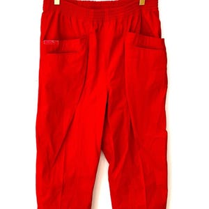 Peut inclure: Pantalon rouge vif avec une taille élastique et deux poches avant. Le pantalon est suspendu à un cintre doré sur fond blanc. Une petite étiquette rectangulaire est visible sur la poche gauche.